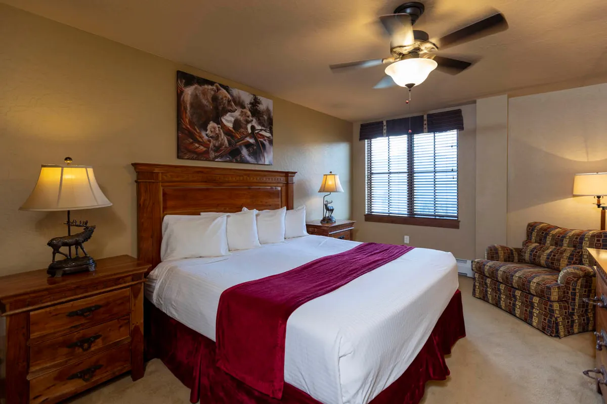 A cozy bedroom featuring a king-sized bed with a vibrant red throw, wooden nightstands, a comfortable armchair by the window, and playful bear-themed artwork. Gentle natural light filters through the blinds, creating a restful and inviting atmosphere.