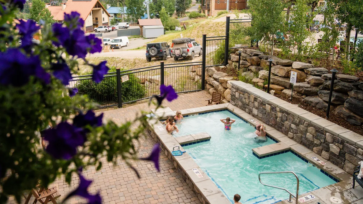 An inviting outdoor resort pool area featuring a sparkling swimming pool, adjacent hot tub, and colorful landscaped surroundings. Lounge chairs invite relaxation, and the setting suggests both recreation and social interaction, possibly with mountain views in the distance.