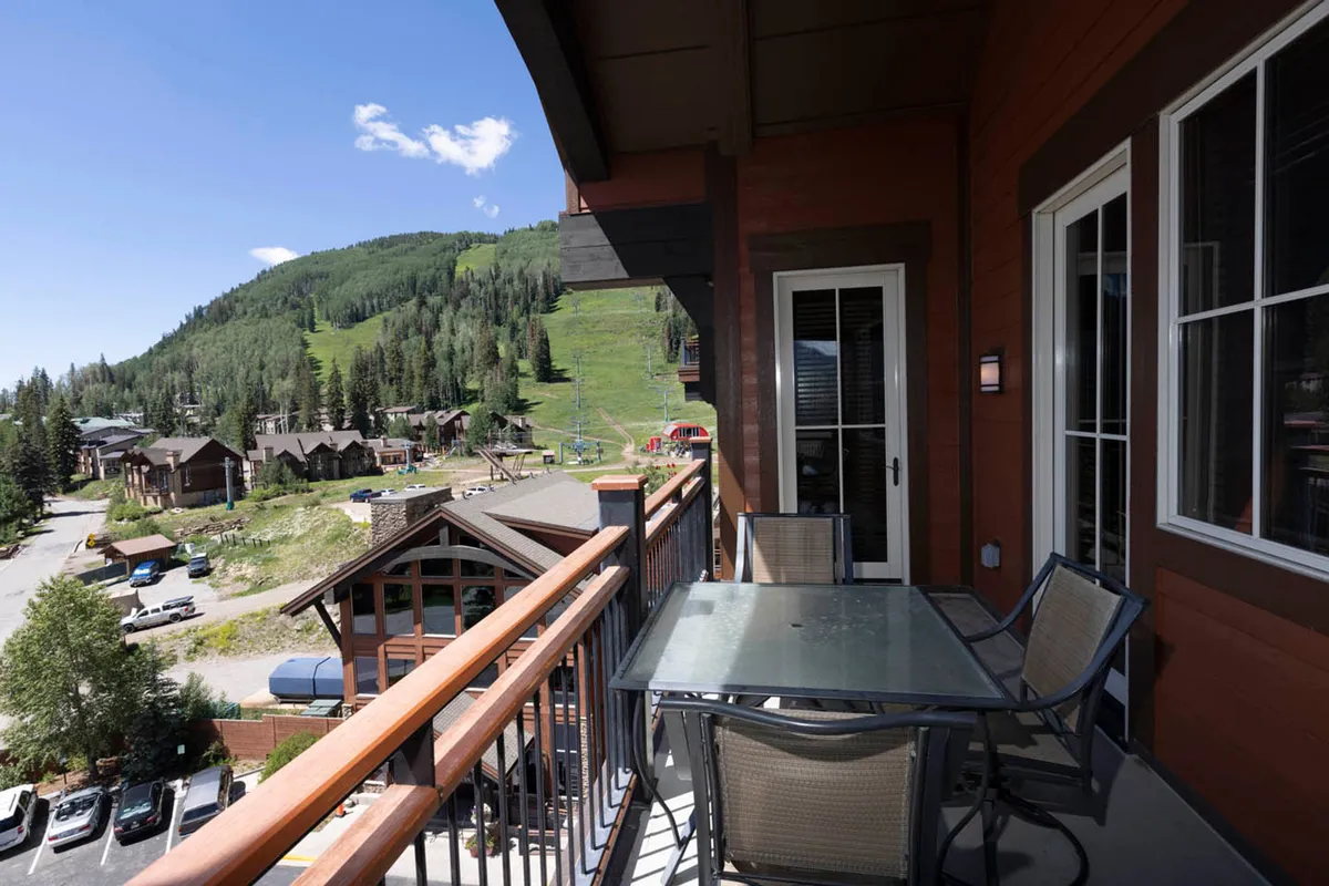 An exterior view from the private balcony, complete with comfortable seating, overlooking the dramatic Needles Mountain Range and nearby village buildings. The scene captures relaxation, outdoor socializing, and the grandeur of the mountain setting.