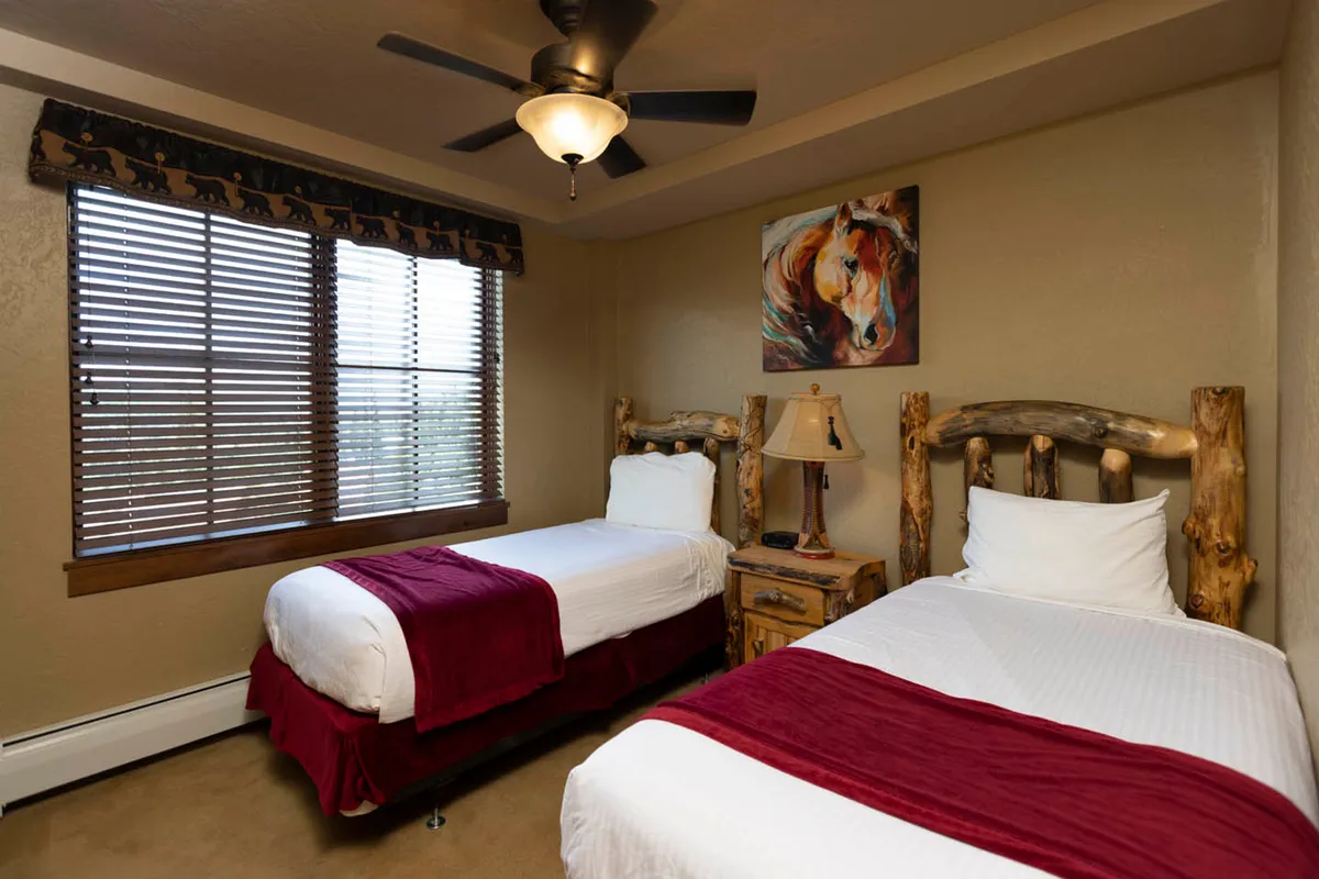 A cozy twin bedroom with two rustic wooden beds, a bedside lamp casting a warm glow, and a painting of a horse. The room feels inviting, perfect for kids or friends, and offers a sense of rustic mountain charm.