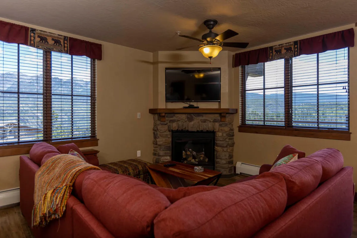 An inviting living space featuring a prominent red couch, a stone fireplace, a flat-screen TV, and large windows. The scene suggests comfort and social activities, such as movie nights or relaxing evenings by the fire, all while enjoying scenic vistas.