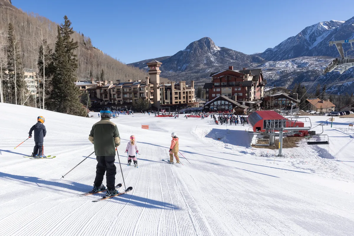 A lively scene with families and friends skiing down a snow-covered slope, with the resort buildings and ski lift visible in the background. The image evokes the joy and togetherness of a mountain getaway.