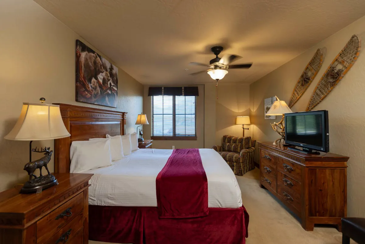 This image depicts a king-sized bed with red accents, complemented by warm wooden furniture, wall artwork, and a window with blinds. The inviting and tranquil ambiance is enhanced by soft lighting and carefully chosen decor.