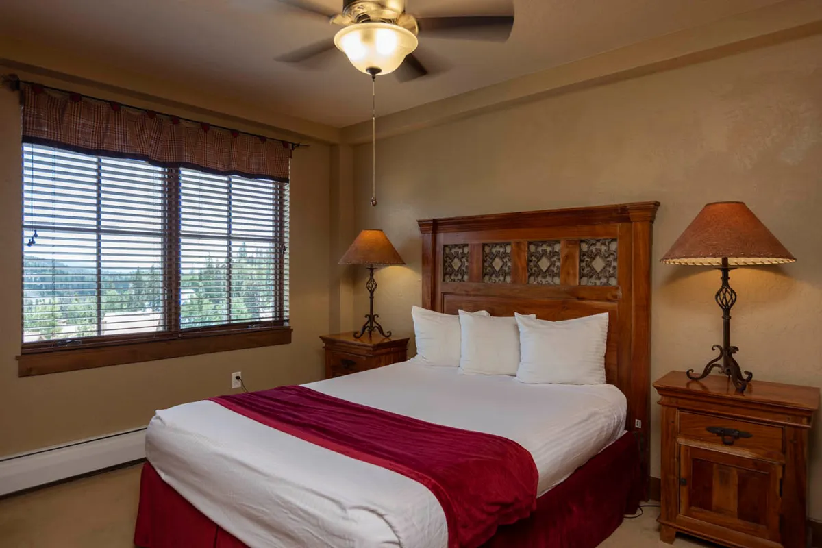 This inviting bedroom features large windows with blinds, allowing guests to enjoy fresh mountain air and natural light. The room is cozy, with white linens, red accents, and wooden nightstands, suggesting comfort and relaxation after a day of adventure.