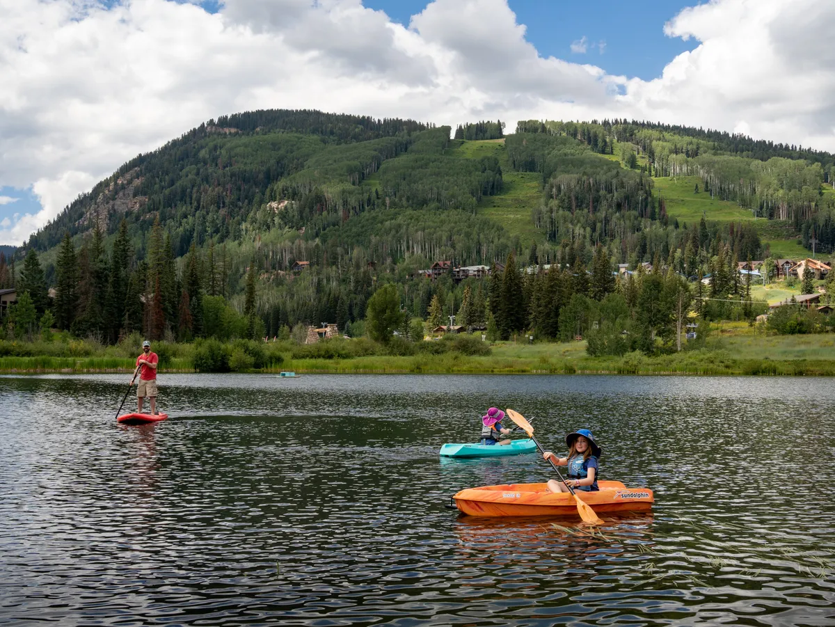 This vibrant scene shows a paddleboarder and kayakers enjoying a serene mountain lake surrounded by dense greenery and towering peaks. The water reflects the sky and trees, emphasizing outdoor activities and the region’s natural splendor.