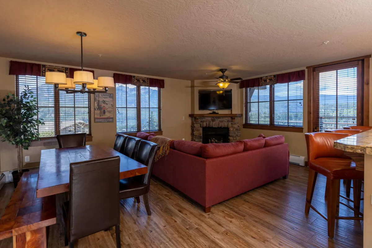 A cozy interior shot showcasing a stone gas fireplace, a comfortable sofa, a dining table, and expansive windows that flood the space with natural light. The room invites relaxation, conversation, and family gatherings, with the windows drawing attention to possible mountain views beyond.