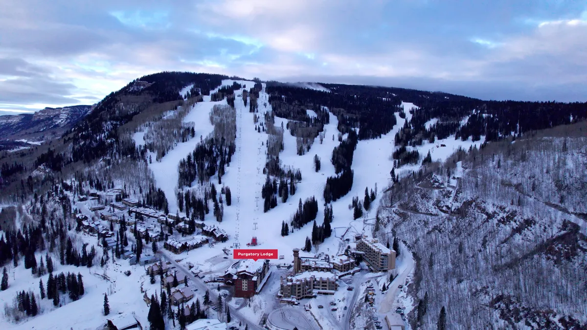 A sweeping exterior shot of Purgatory Lodge nestled at the base of snow-covered ski slopes, with mountains rising in the background. The scene captures skiers and snowboarders in action, the lodge’s proximity to the slopes, and the picturesque natural setting.