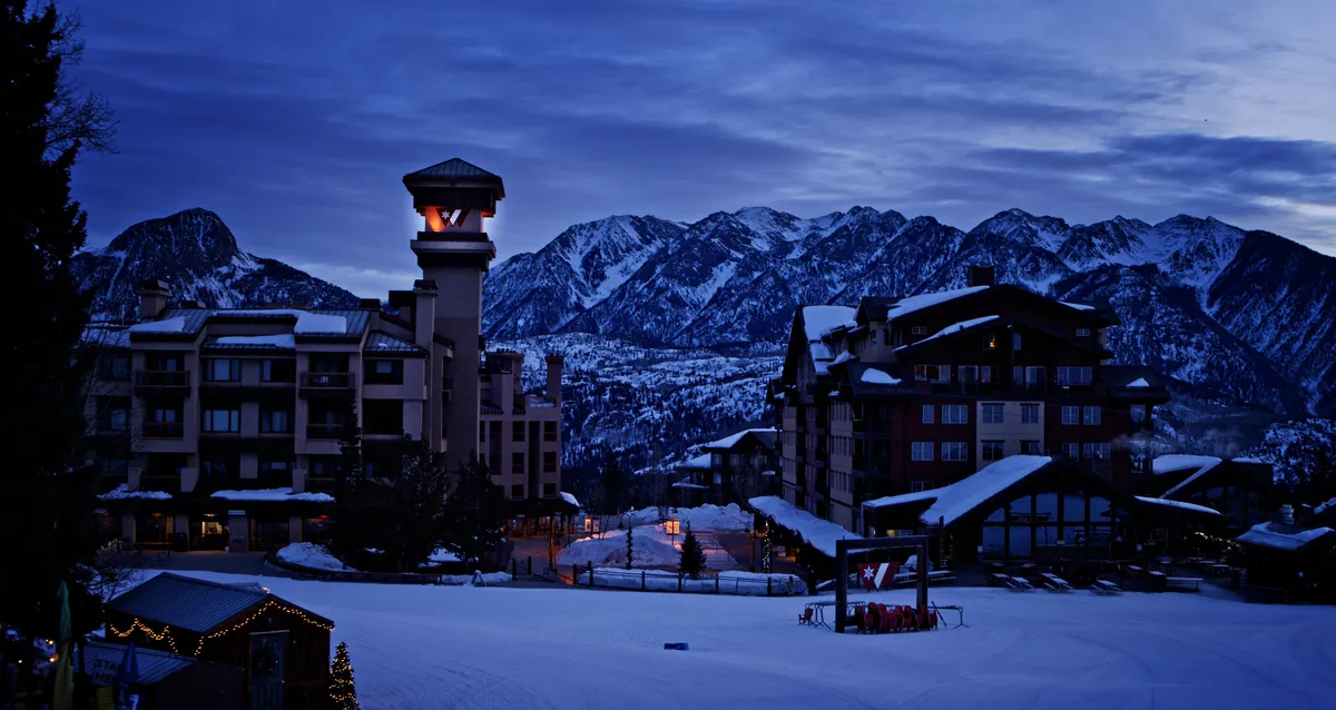 An inviting exterior view featuring a majestic mountain range as the backdrop to the resort’s charming architecture. The snow-covered ground, illuminated tower, and clear mountain air evoke a sense of serenity and connection to nature—key elements of the wellness and relaxation narrative. The image hints at outdoor activities and peaceful moments like watching sunsets or stargazing on the balcony.